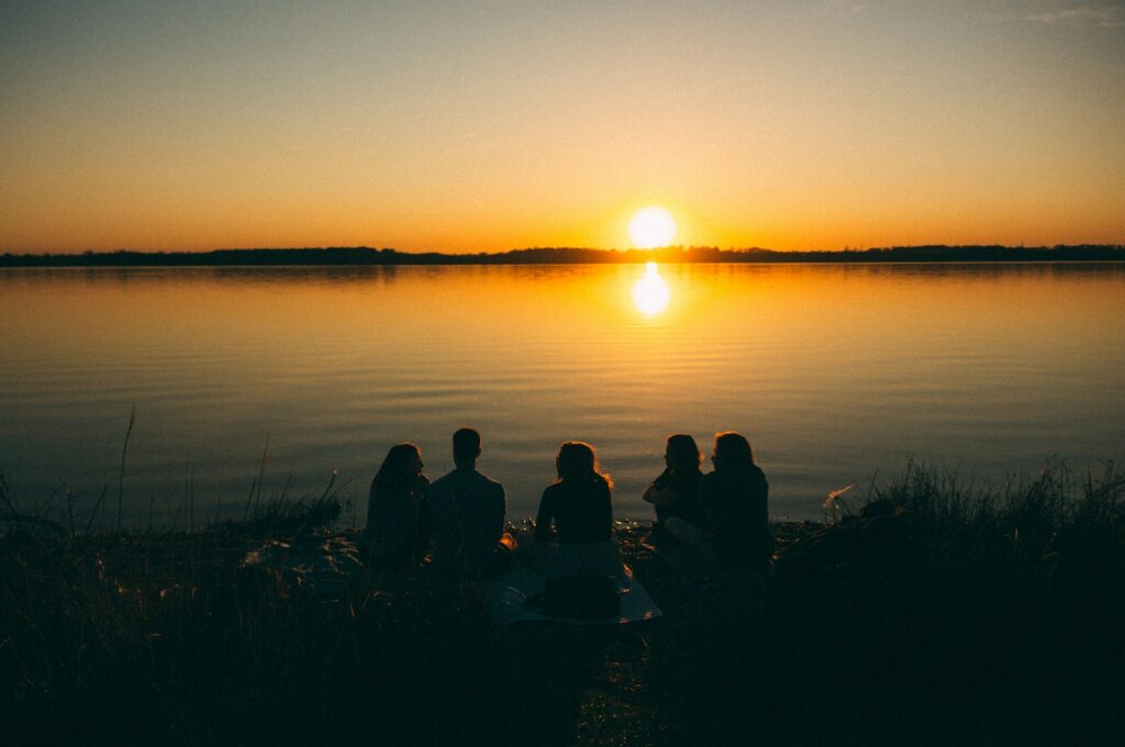 a group of people sitting on the shore of a lake at sunset